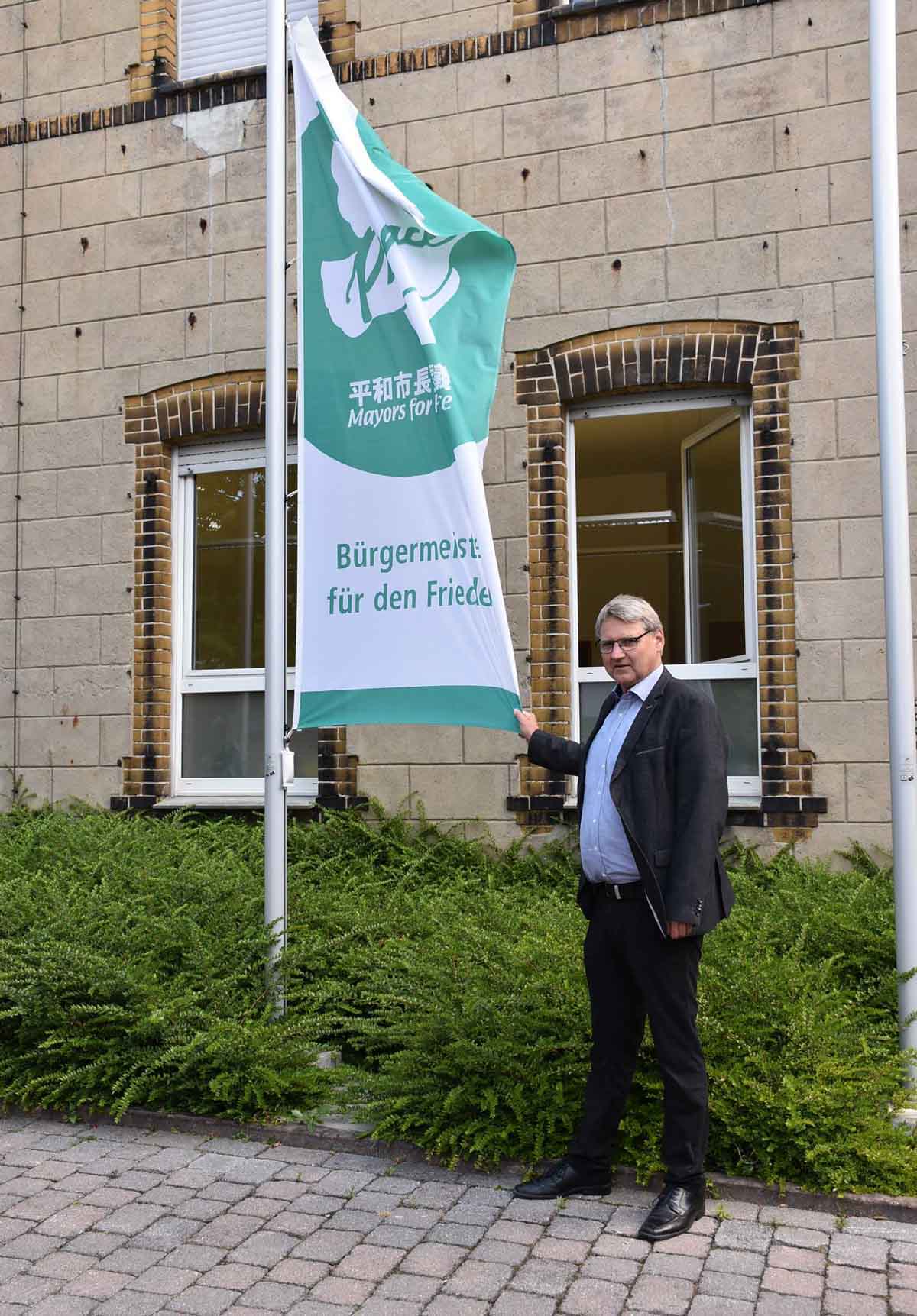 Klaus van Roje setzt als allgemeiner Vertreter des Bürgermeisters ab dem 8. Juli mit der Flagge der Mayors for Peace am Rathaus in Ascheberg wieder ein Zeichen für den Frieden und gegen Atomwaffen. Foto: Gemeinde Ascheberg