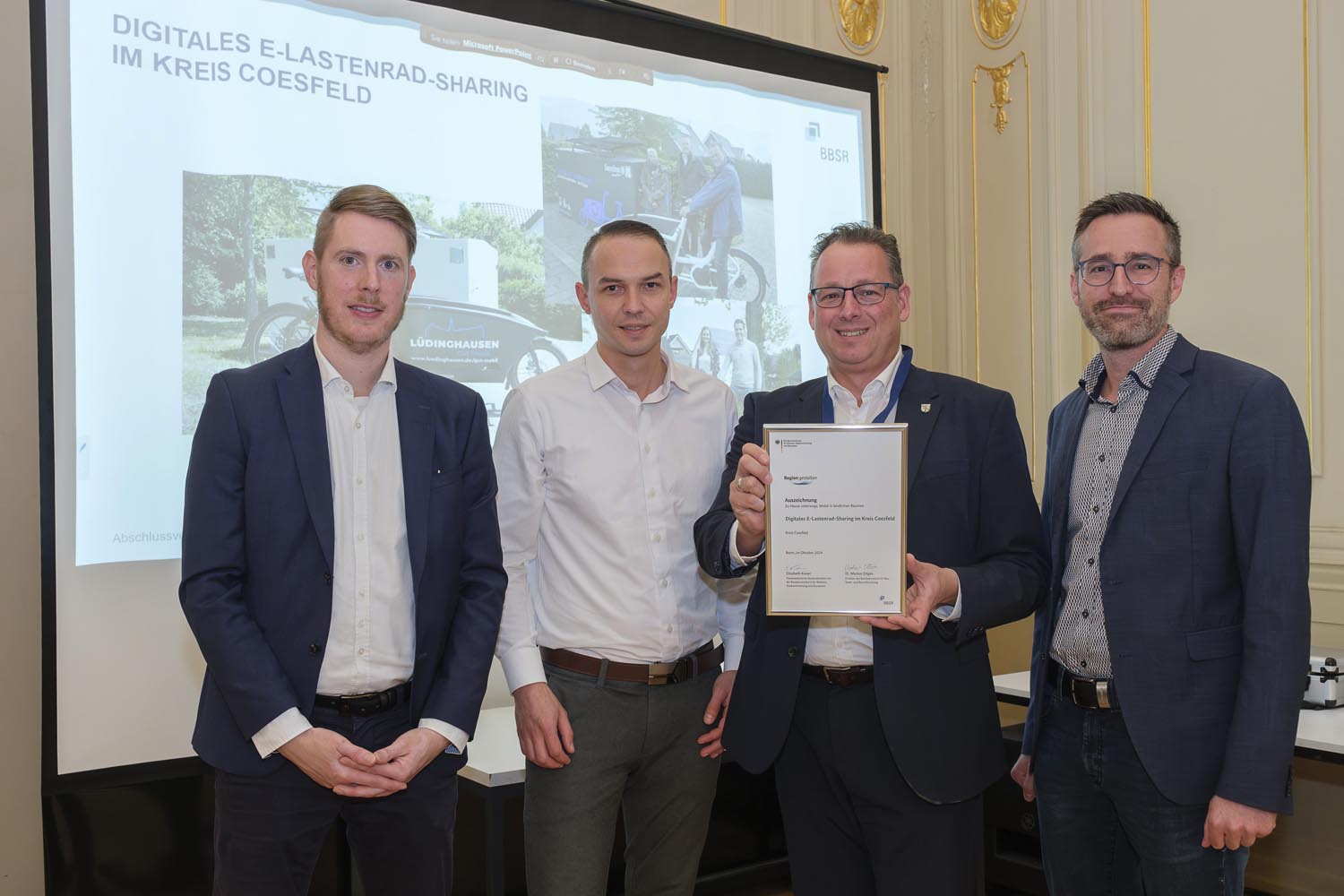 Nahmen am 24. Oktober stellvertretend für die beteiligten Kommunen die Siegerurkunde von Johannes Schneider (l.) vom Kompetenzzentrum für Ländliche Mobilität entgegen: Niklas Esser und Bürgermeister Sebastian Täger (Gemeinde Senden) und Mathias Raabe (Sprecher der Digitalagentur Kreis Coesfeld). Foto: BBR, Bundesfoto, Guido Schiefer